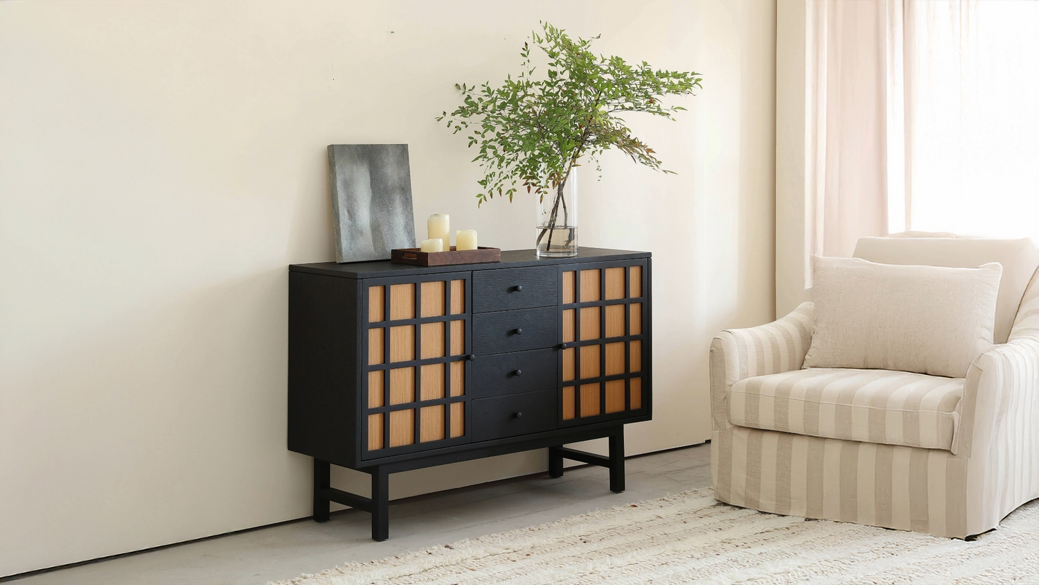 Black wood sideboard with grid-paneled natural oak doors, featuring minimalist candle decor, a tall glass vase with green branches, and a striped accent chair in a bright modern living room.