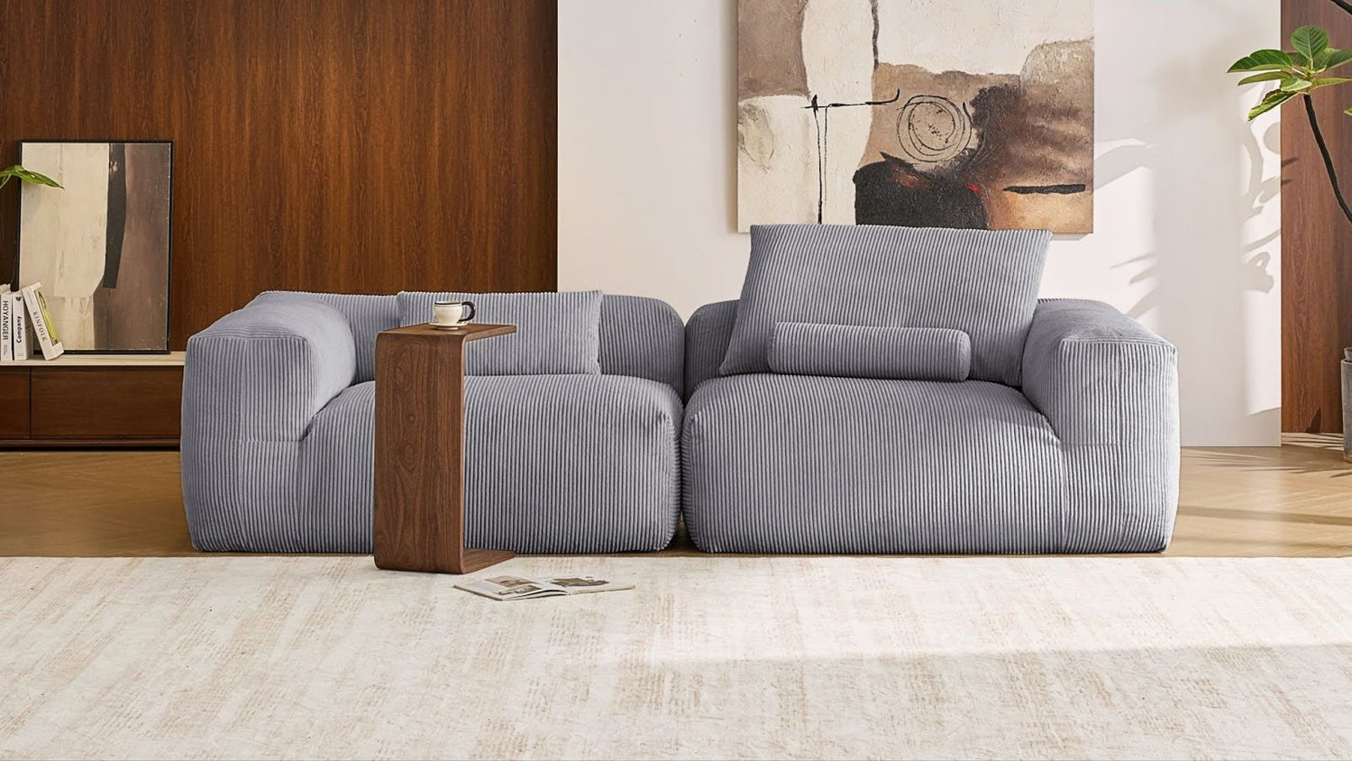 Two-seat modular corduroy sofa in slate blue with low-profile, plush seating and a wood wraparound side table, styled against a walnut-paneled wall—perfect for compact living rooms seeking comfortable furniture with built-in surface functionality.